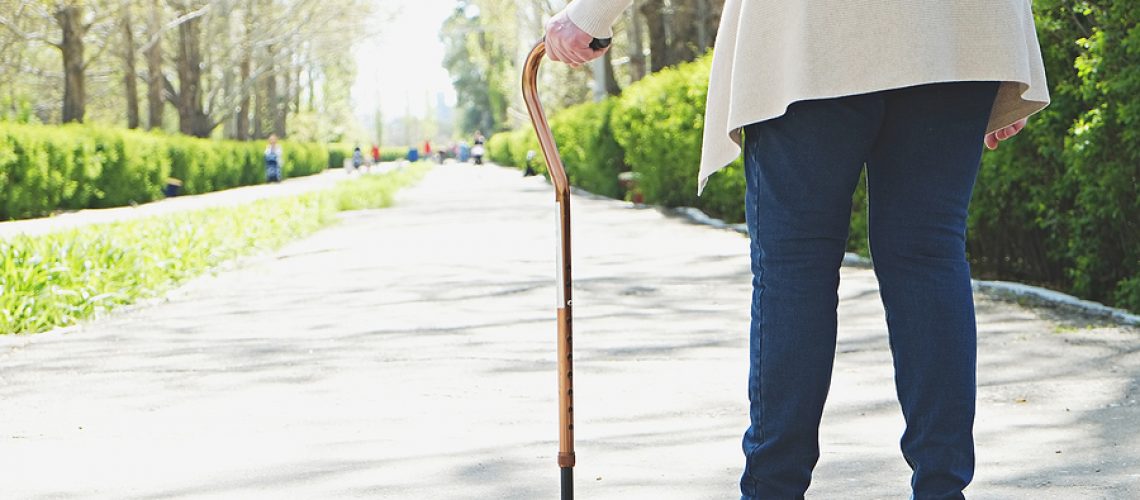 Senior disabled caucasian woman hands on cane outside nursing home park. Close up of elderly lady holding a walking stick outdoors of healthcare facility on the sunny day.